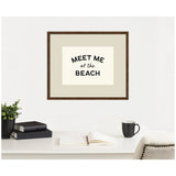 Framed wall art with the text 'MEET ME at the BEACH' in black on cream background, hung above a white desk with a plant, books, open notebook, and black mug.