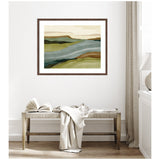 Abstract landscape wall art with layered horizontal shapes in muted green, brown, and gray tones framed in dark wood, hanging above a rustic bench with draped fabric and open book.