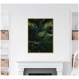 Framed aerial photograph showing a narrow, winding river cutting through a densely green valley with dark forest patches and varying green shades, hung above a white fireplace.