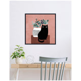 Framed canvas print of a stylized black cat with wide eyes sitting beside a white patterned vase with green foliage on a muted pink background, displayed above a wooden desk with a plant and metal decor.