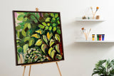 Square canvas painting on an easel depicting green and yellow leafy foliage in an impressionist style with a dark brown frame, set against a white wall with art supplies on shelves nearby.