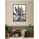 Black and white framed print of tall palm fronds displayed above a wooden bench with a beige pillow and draped blanket, surrounded by potted plants against a two-tone beige and sage green wall.
