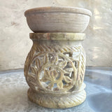 Carved soapstone essential oil burner in light beige with leaf and swirl patterns, topped with a shallow round bowl, placed on a reflective surface.
