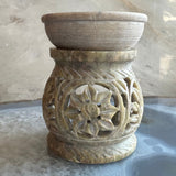 Carved soapstone essential oil burner with a circular top dish and floral openwork pattern on the rounded base, in varied beige and green tones.