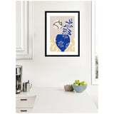 Framed wall art print with a blue vase holding a leafy branch against beige and cream abstract shapes, displayed above a white kitchen countertop with green apples.