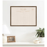 Framed beige linen fabric pinboard printed with vintage postcard back design, hanging on white wall above white surface with lamp, photo frame, plant, and bowl.