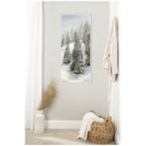 Vertical canvas painting of snow-covered evergreen trees in a muted gray and white winter landscape, hung on a light gray wall above a wicker basket and decorative vase.