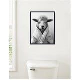 Framed black and white photo of a sheep wearing a plush bathrobe, hung on a white wall above a white toilet tank in a minimalist bathroom.