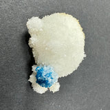 Small, bright blue cavansite crystal ball sitting on larger, white stilbite crystal cluster with rough, sparkling surfaces on dark background