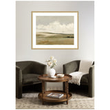 Framed landscape painting of soft beige and gray rolling hills with fluffy white clouds above, hung on a light wall between two dark upholstered chairs and a round wooden table.