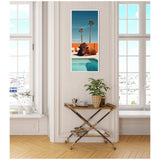 Vertical framed artwork showing a brown bison lying beside a blue swimming pool under a vibrant blue sky with two tall palm trees and an orange wall in the background, hung on a white paneled wall between two large windows.