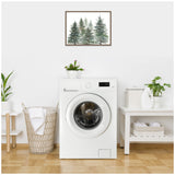 Framed watercolor painting of pine and evergreen trees in green and blue hues hanging above a white front-loading washing machine in a bright laundry room with plants, a wicker basket, and folded linens.