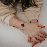 Close-up of a wrist wearing a thin black heart-shaped macrame bracelet and a delicate gold chain bracelet on a beige ribbed sweater sleeve.