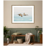 Framed wall art print of two cranes flying against a soft blue and beige sky, hung above a wooden bench with a beige cushion and draped throw in a muted green and beige room.