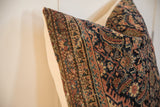 Close-up of a 24x24 pillow featuring an intricate vintage rug fragment pattern with earthy tones, dark blue accents, and worn texture on a light background.