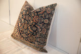 Square pillow with intricate vintage rug-inspired pattern in dark blue, red, beige, and green tones resting against a white wall on a light-colored fabric surface.