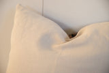 Close-up of a cream-colored square pillow with a textured fabric surface and soft folds against a pale background