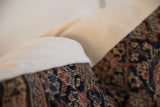 Close-up of a vintage rug fragment pillow showing intricate red, blue, and beige geometric patterns next to a plain beige fabric edge.
