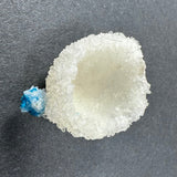 White crystalline Stilbite formation with a porous, rounded structure and a small bright blue cavansite ball attached on the side, displayed on a gray background.