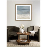 Framed abstract wall art in shades of blue and beige above two dark olive armchairs with a round wooden coffee table holding a white vase with flowers and small bowls.