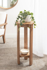 Round wooden side table with tall legs and a lower shelf holding a ceramic bowl, topped with a white pot containing a trailing green plant against a bright window.