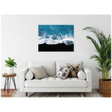 Framed aerial photograph of ocean waves crashing on a black sand beach, hung above a white sofa with neutral and patterned throw pillows in a minimal living room.