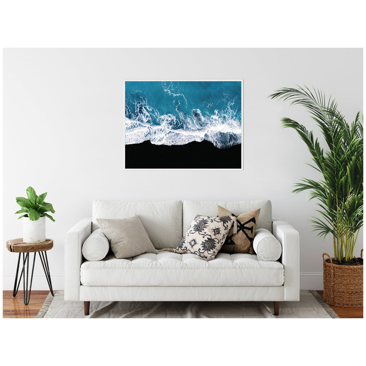 Framed aerial photograph of ocean waves crashing on a black sand beach, hung above a white sofa with neutral and patterned throw pillows in a minimal living room.