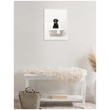 Framed canvas of a black Labrador sitting in a white clawfoot bathtub filled with bubbles, mounted on a light neutral wall above a white bench with a fluffy throw and beige bag below.