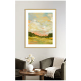 Framed impressionist landscape painting featuring a pastel sky with soft clouds above a green field bordered by dark trees in a modern living room setting.