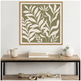 Square canvas artwork with white botanical leaf silhouettes on an olive green background, framed in light wood, hanging above a rustic wood and black metal console table with neutral decor.