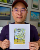 Person holding a watercolor painting titled 'Biking in the Light' showing cyclists on a tree-lined path with dappled sunlight and a distant blue river horizon.