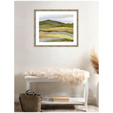 Abstract landscape artwork featuring layered hills in muted greens and browns, framed and hung on a white wall above a white bench with a beige fur throw.