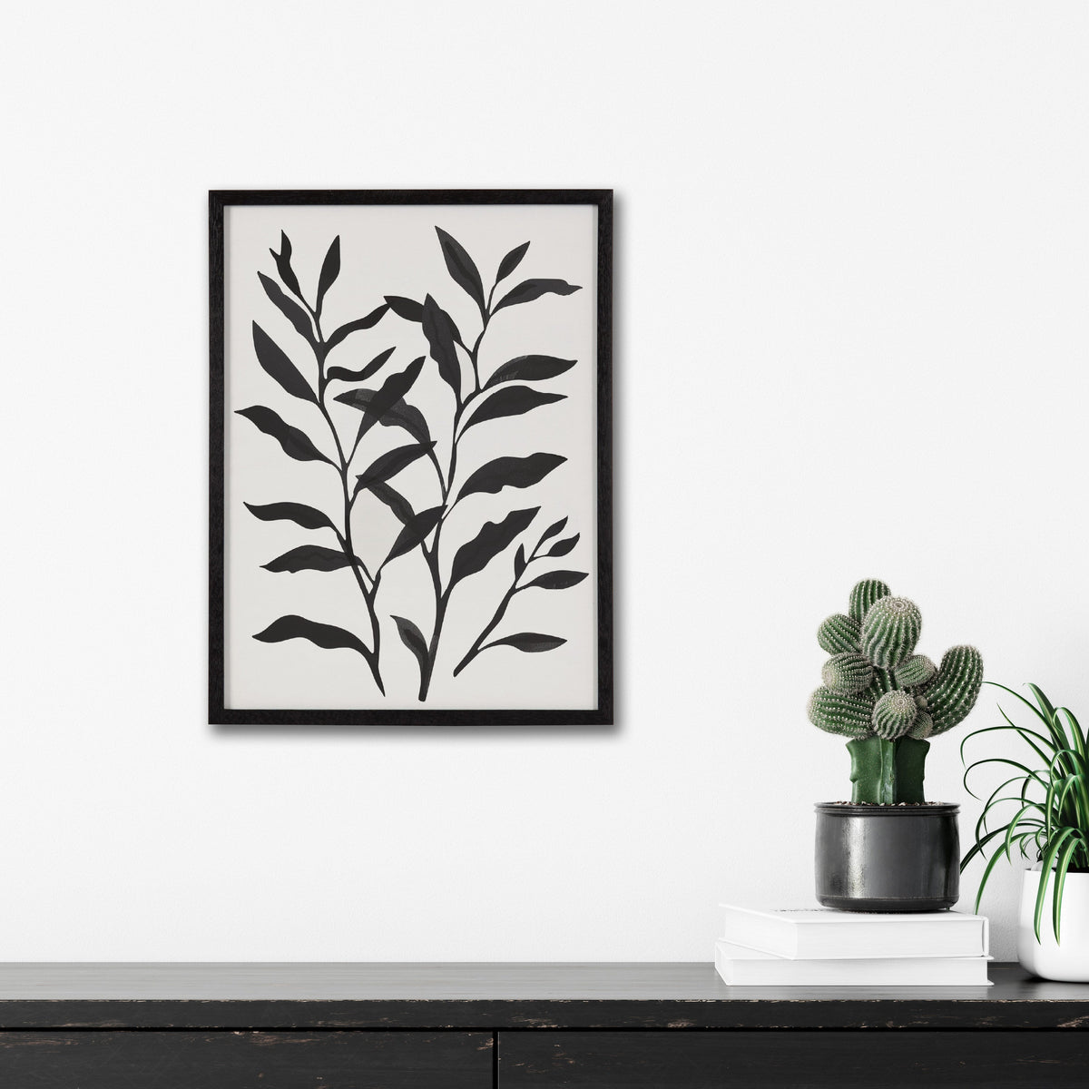 Black silhouette of botanical leaves with elongated shapes on a white background framed in black, hanging above a dark wood surface with potted cacti and books.
