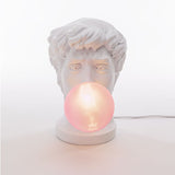 White sculpted bust resembling a classical male head with tousled hair, holding a glowing pink orb at its mouth on a round base with visible power cord
