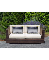 Two-seater dark brown wicker outdoor sofa with beige cushions and two rectangular blue and white striped decorative pillows, set on a gray stone patio with green shrubbery in background.