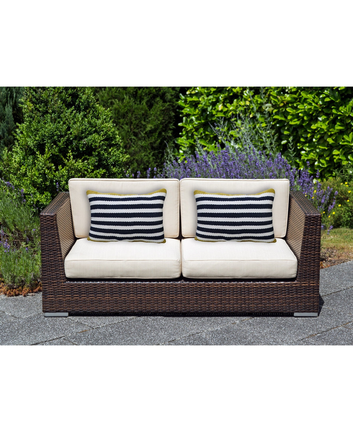Two-seater dark brown wicker outdoor sofa with beige cushions and two rectangular blue and white striped decorative pillows, set on a gray stone patio with green shrubbery in background.