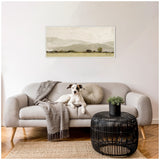 Neutral-toned abstract landscape painting with textured brushstrokes depicting hills and trees, hanging above a modern beige sofa with a dog, knit blanket, and round cushion.
