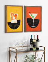 Two framed prints showing stylized cocktails, one with an orange background and ice cubes, the other with a red background and olives, above a gold-framed glass bar cart with wine bottles and glasses.