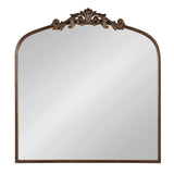 A rectangular mirror with a rounded arched top and an ornate dark bronze decorative crest featuring carved scrollwork at the center of the arch.