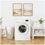Minimalist laundry room with white front-load washing machine between a wicker laundry basket and a white table holding white detergent containers and a small plant, two pastel landscape paintings above.
