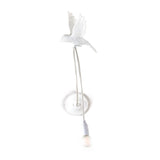 White wall lamp shaped like a flying sparrow with an exposed bulb hanging from a flexible cord below, mounted on a round base.