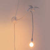 White wall lamp featuring a bird figure perched on a curved arm holding a glowing bulb, casting soft warm light and shadows on a muted wall.