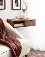 Wall-mounted wooden side table with a drawer and open shelf bearing light-colored books, topped with white vases and a textured glass candle holder beside a cream armchair and rust-colored knit blanket.