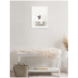 Framed canvas showing a black and white French Bulldog sitting in a white clawfoot bathtub, hung on a light gray wall above a white bench with a beige fur throw and a gray bag beside it.
