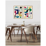 Two framed abstract canvases with colorful geometric shapes in blue, green, red, and yellow hang above a modern wooden dining table with cream chairs on light wood flooring.