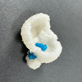 White, crystalline stilbite formation with two bright blue cavansite crystal balls nestled within on a dark gray surface.