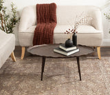 Oval coffee table with a wavy edge and three slender legs, finished in dark brown, placed on a patterned beige rug in front of a cream sofa with a rust-colored knit throw.