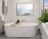 Minimalist bathroom featuring a white freestanding oval bathtub with wooden tray holding a plant, beneath a framed ocean wave print on a neutral wall.