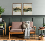 Two framed abstract landscape canvases with muted green, beige, and black tones hanging above a wooden sofa with dusty pink cushions and a round beige pillow