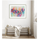 Abstract horizontal painting with multicolored vertical brushstrokes in vibrant pinks, blues, oranges, and greens framed in light wood, hanging above a beige bench with draped fabric.